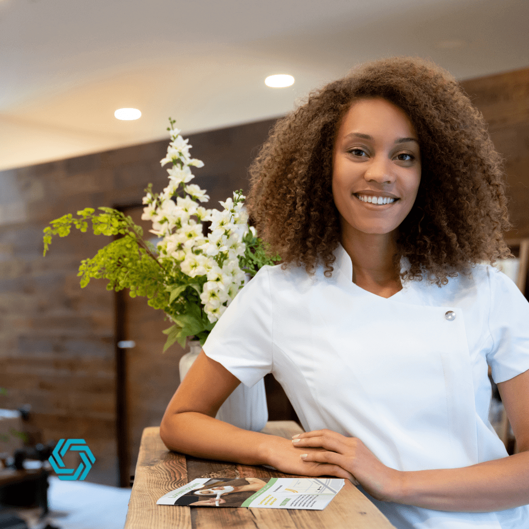 Smiling Woman In White T-shirt Sitting At A Table With A Plant In The Background.